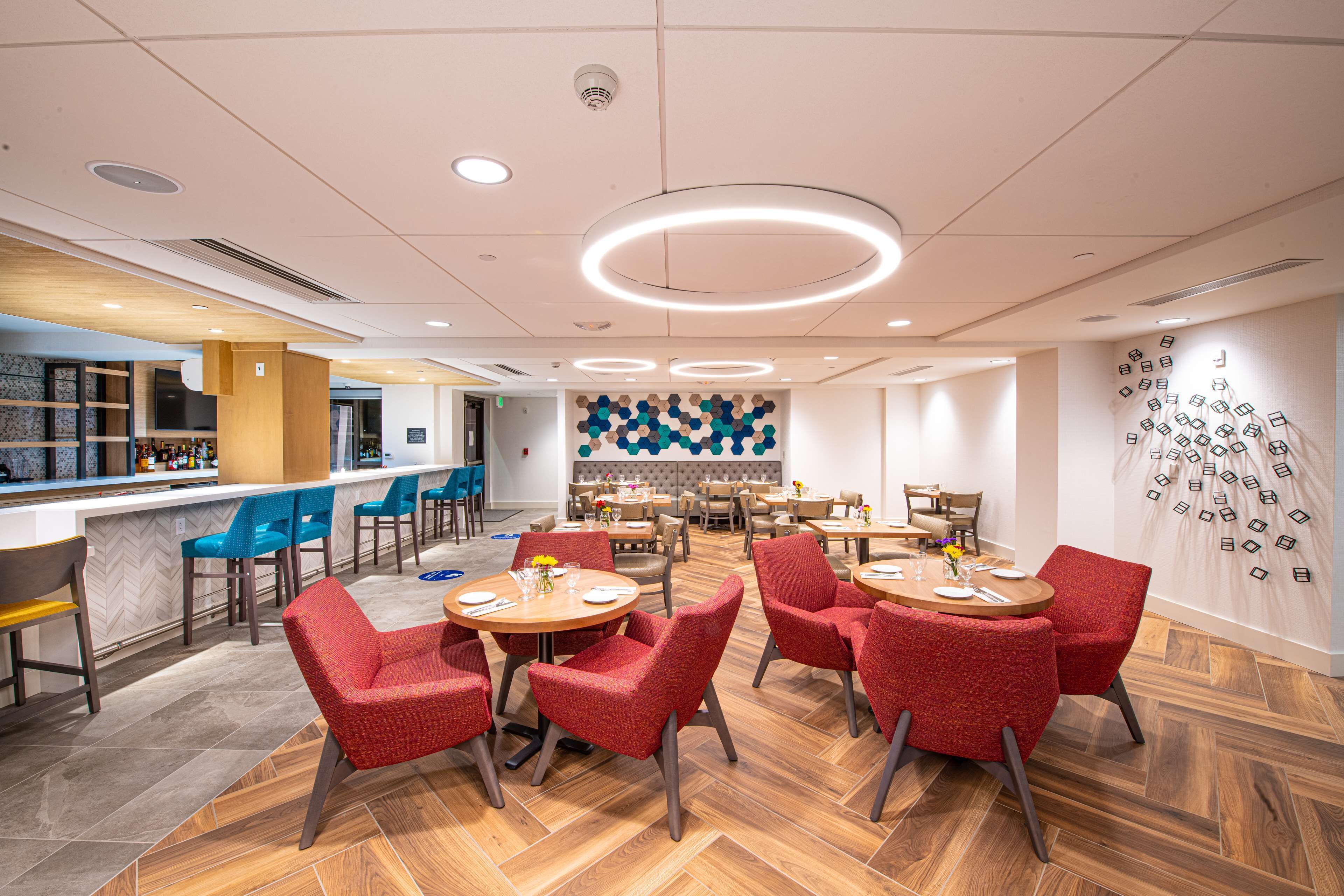 A modern restaurant with round tables and red chairs, a bar area, and a decorative wall with hanging elements.