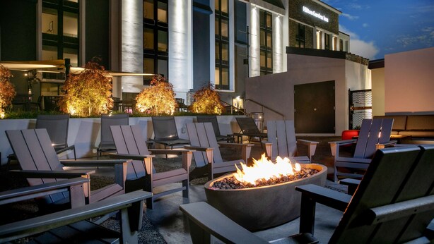 Un área de estar al aire libre con sillas y un pozo de fuego, un edificio moderno con un letrero que dice "Hilton Garden Inn" al fondo.