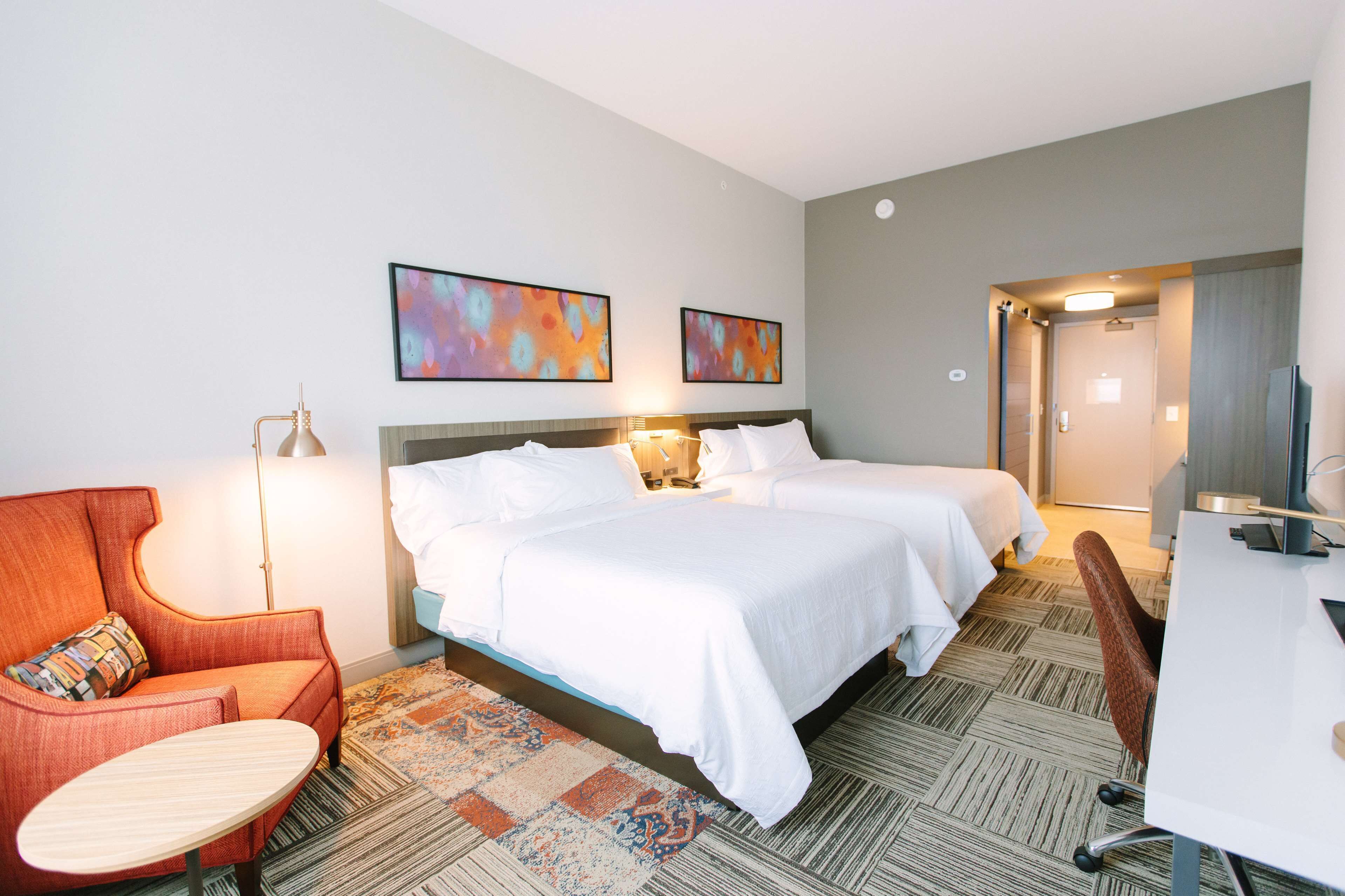 A hotel room with two beds, a desk, a chair, a lamp, and two abstract paintings on the wall.