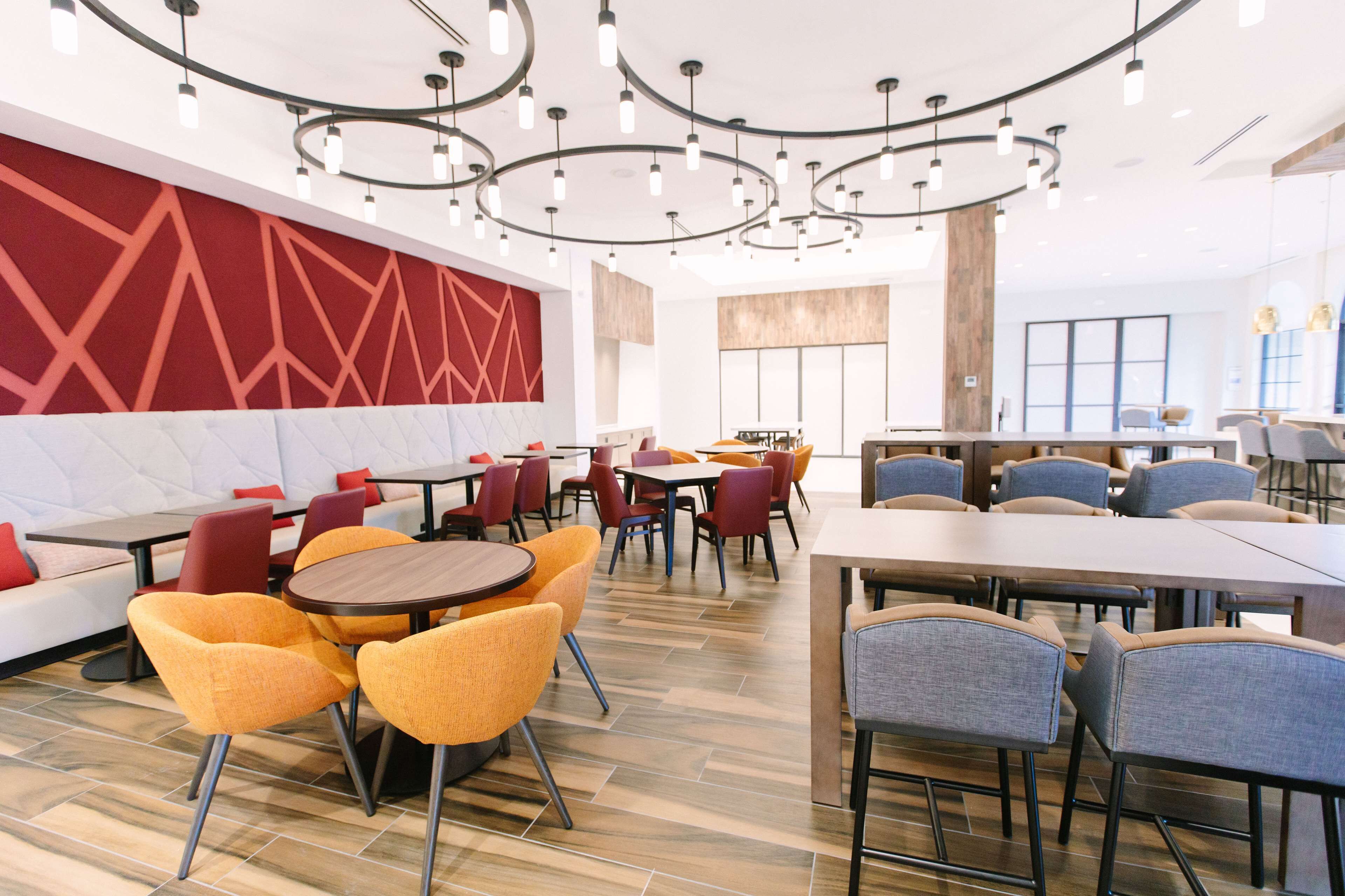 A modern restaurant with round tables and colorful chairs, a red geometric wall, and a wooden floor.