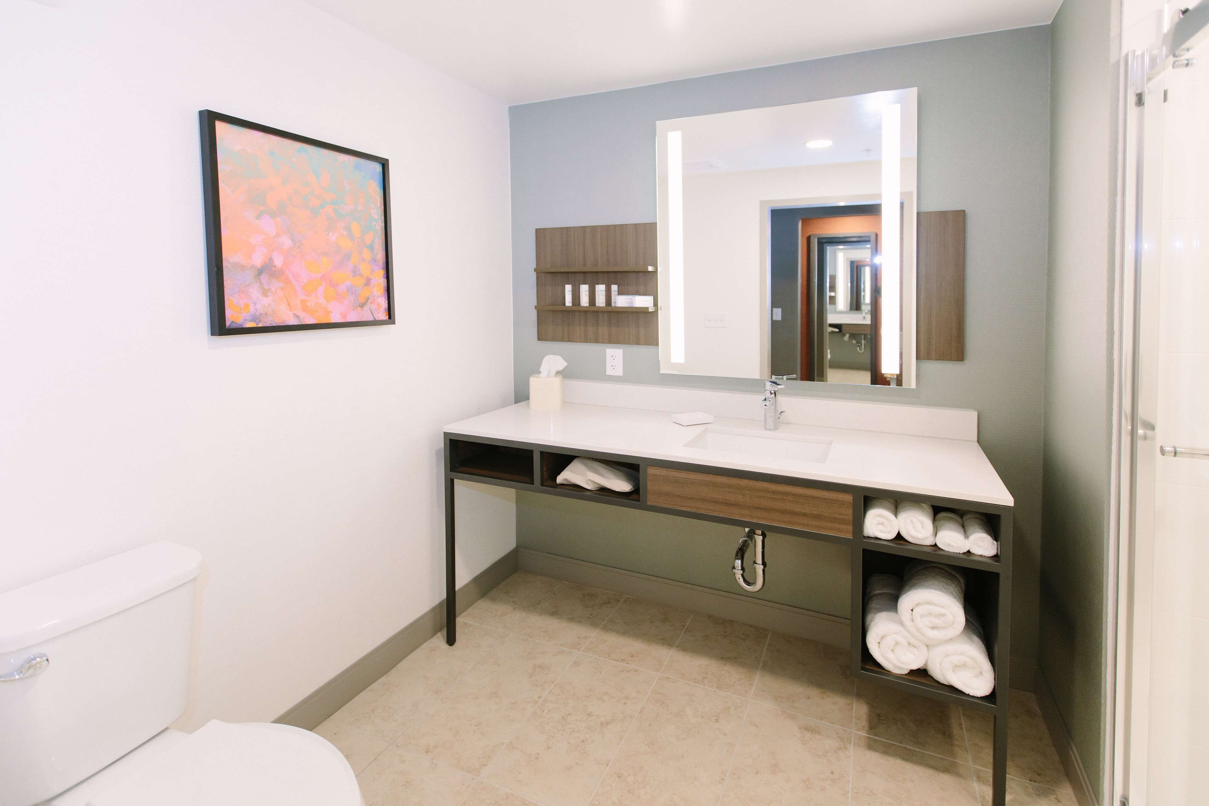 A modern bathroom with a white toilet, a large mirror, a sink with a wooden vanity, and shelves with towels.