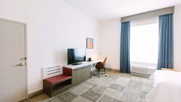 A hotel room with a bed, a desk with a chair, a television, and a window with curtains.