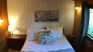 Et soveværelse med seng, puder, et natbord, en lampe, et billede på væggen og et vindue med gardiner.