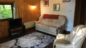 Et hyggeligt rum med træloft, en hvid sofa, en hvid lænestol, et fladskærms-tv og en tæppe med et mønster.
