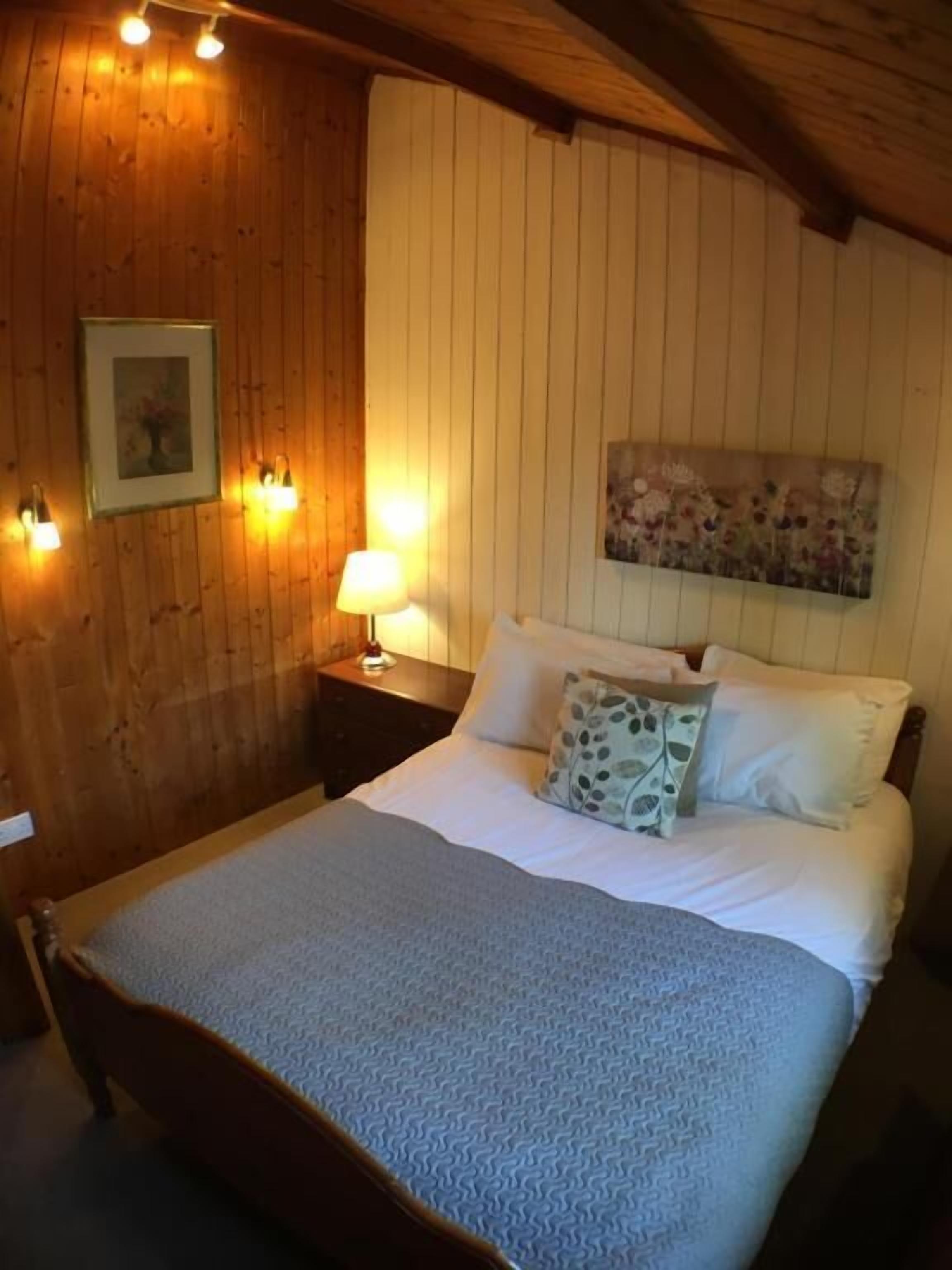 A bedroom with a wooden ceiling, a bed with white bedding, a nightstand with a lamp, and a floral painting on the wall.