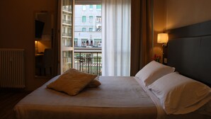 Habitación de hotel con cama, mesita de noche, lámpara y vistas a un balcón con edificios.