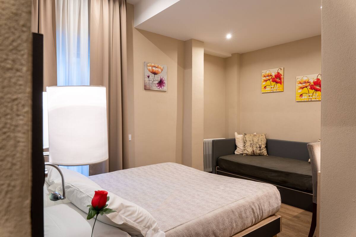 A hotel room with a bed, a bedside table with a lamp, a chair, and two paintings on the wall.