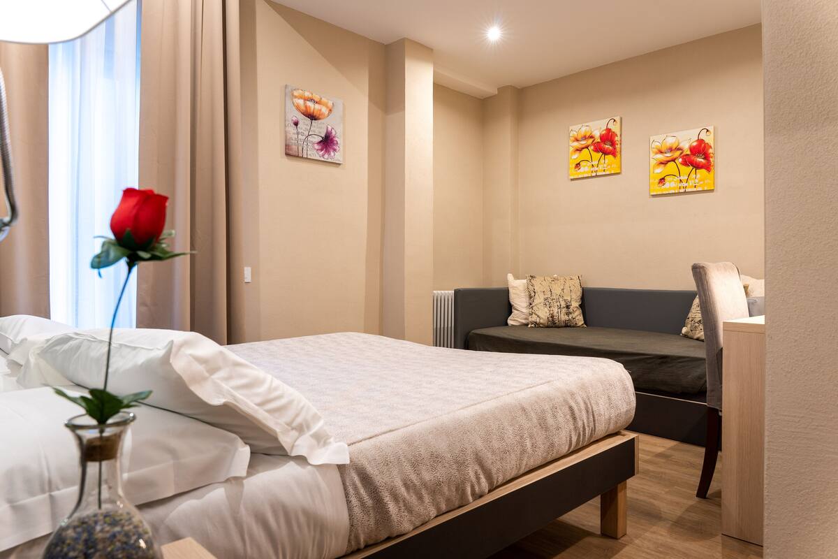 A hotel room with a bed, a bedside table, a chair, a small table, and a vase with a rose.