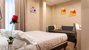 A hotel room with a bed, a bedside table, a chair, a small table, and a vase with a rose.