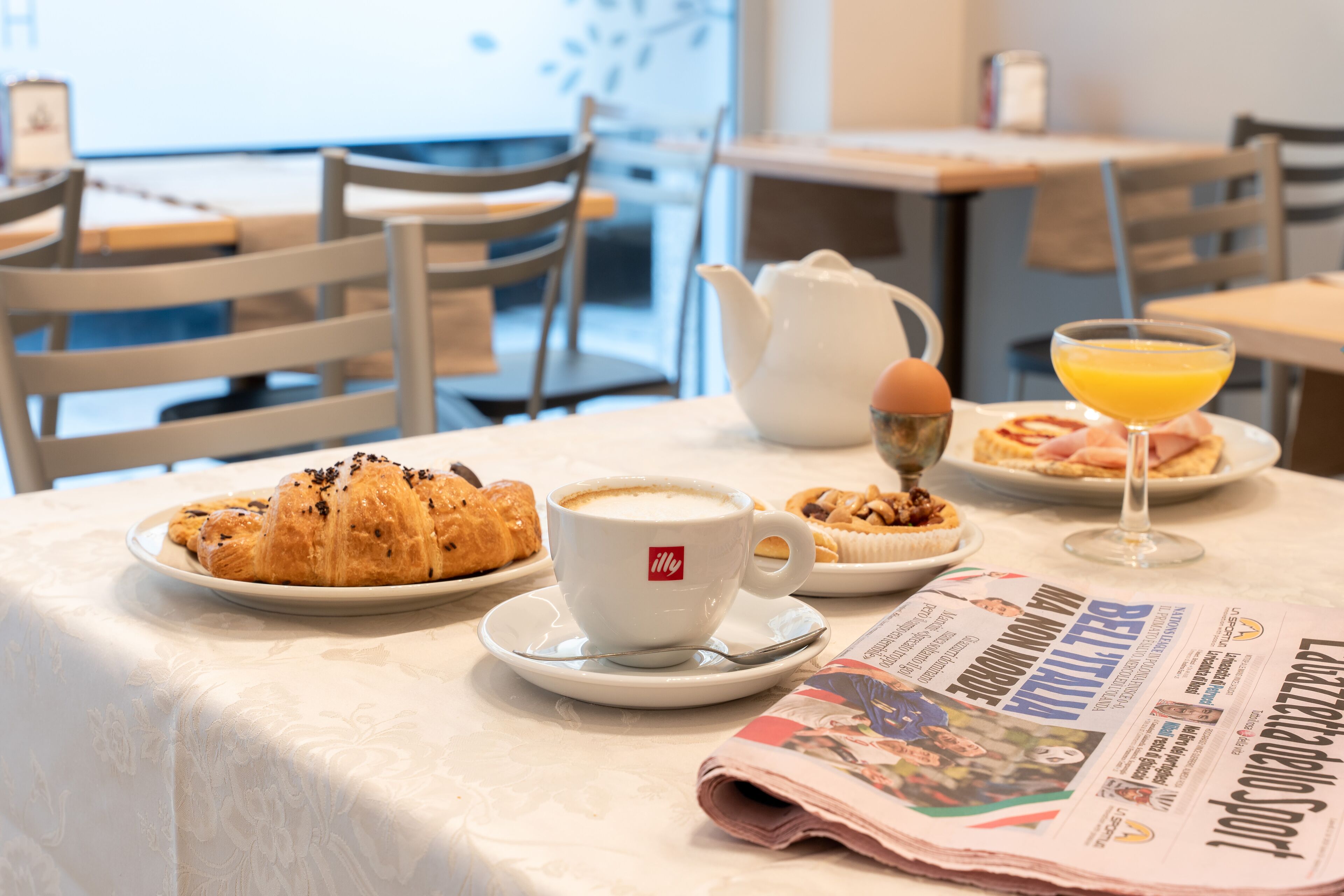 Una mesa de desayuno con croissants, una taza de café, un vaso de jugo de naranja, un periódico y una tetera.