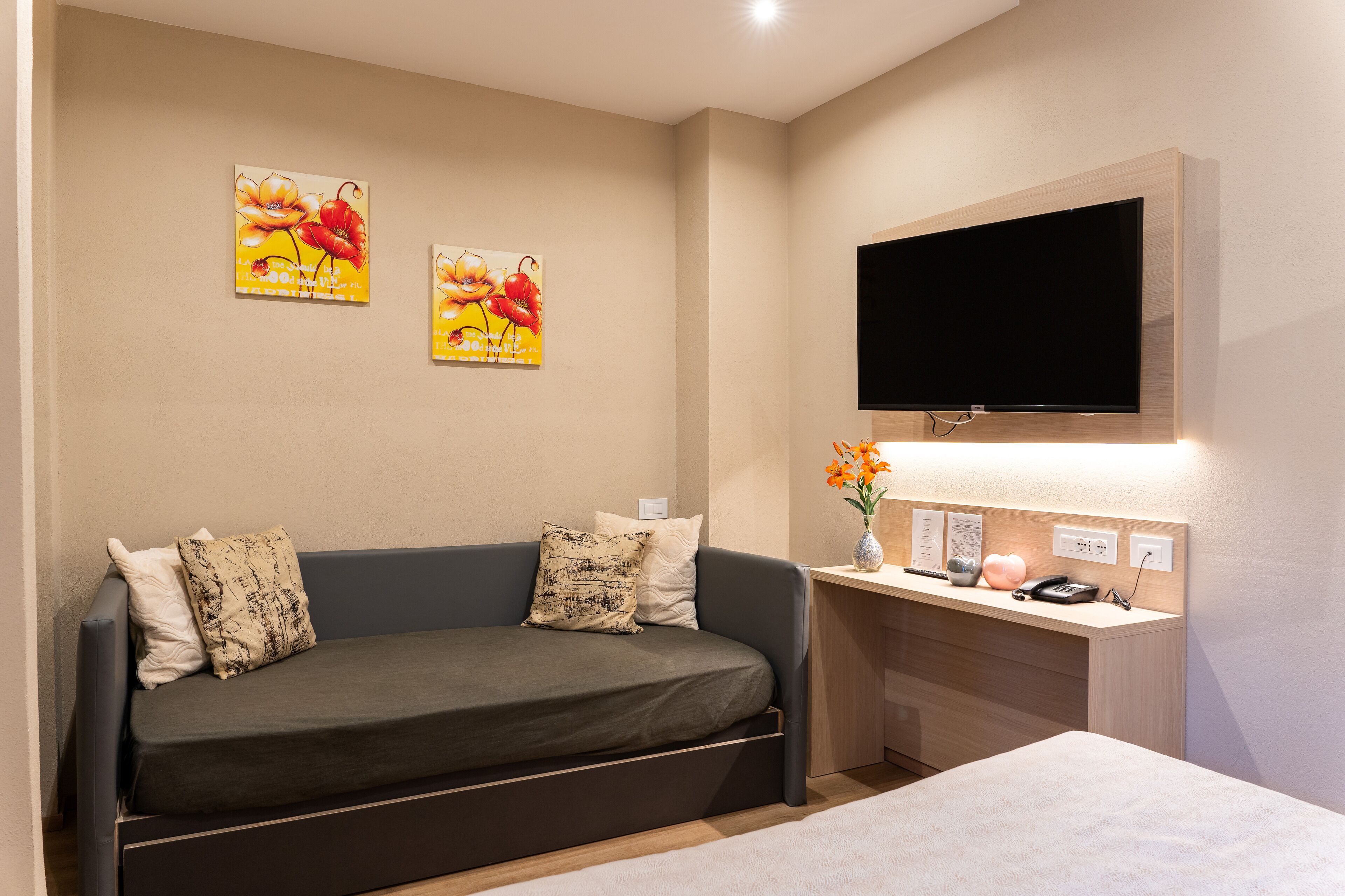 A modern room with a sofa, a flat-screen TV mounted on the wall, and a desk with a vase of flowers.