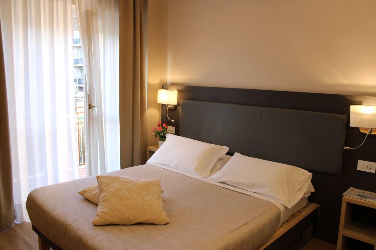 A hotel room with a bed, a nightstand, a lamp, and a window with curtains.
