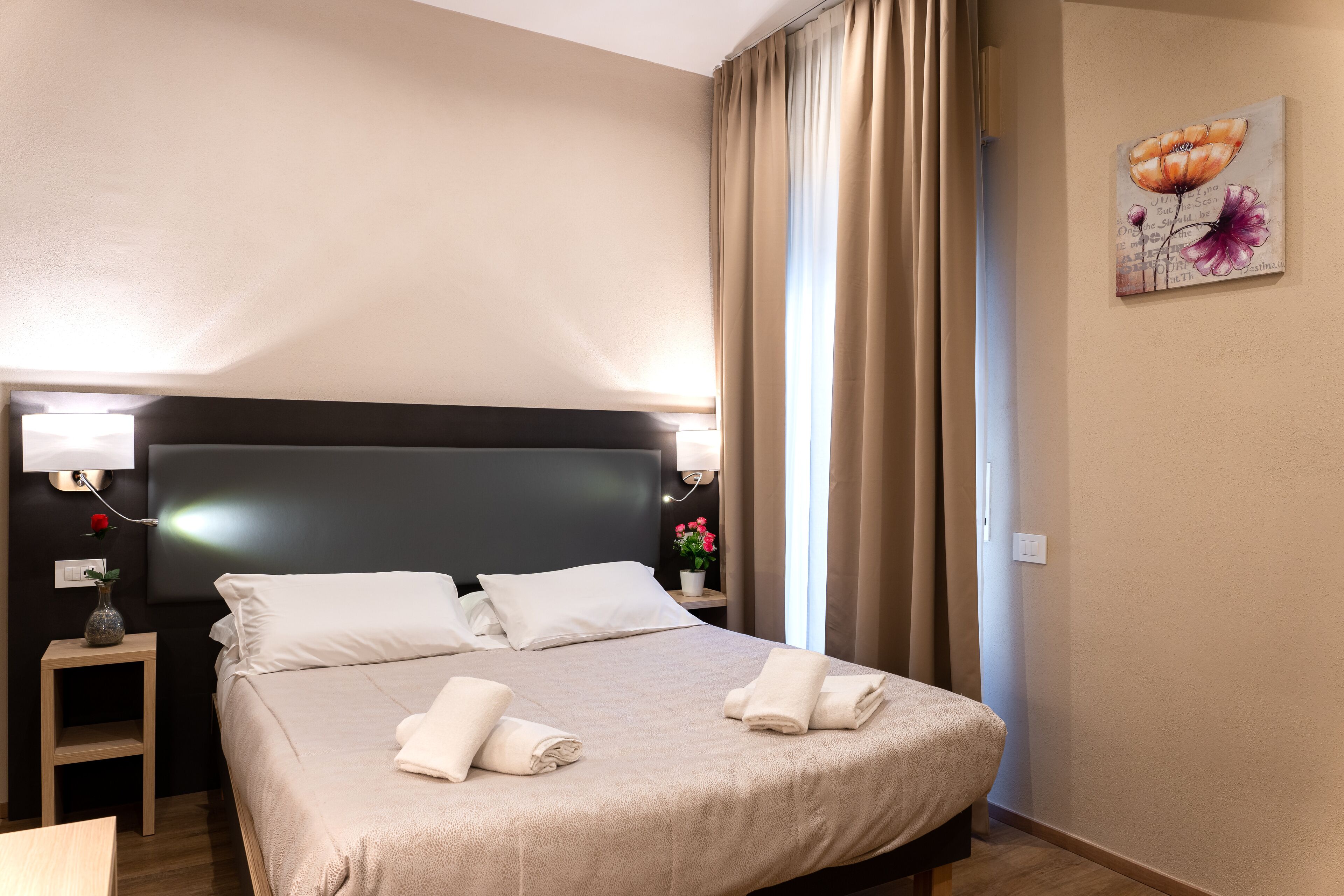 Un lit bien fait, avec du linge de lit et des serviettes blanches, une table de chevet avec une lampe et une œuvre d’art accrochée au mur.