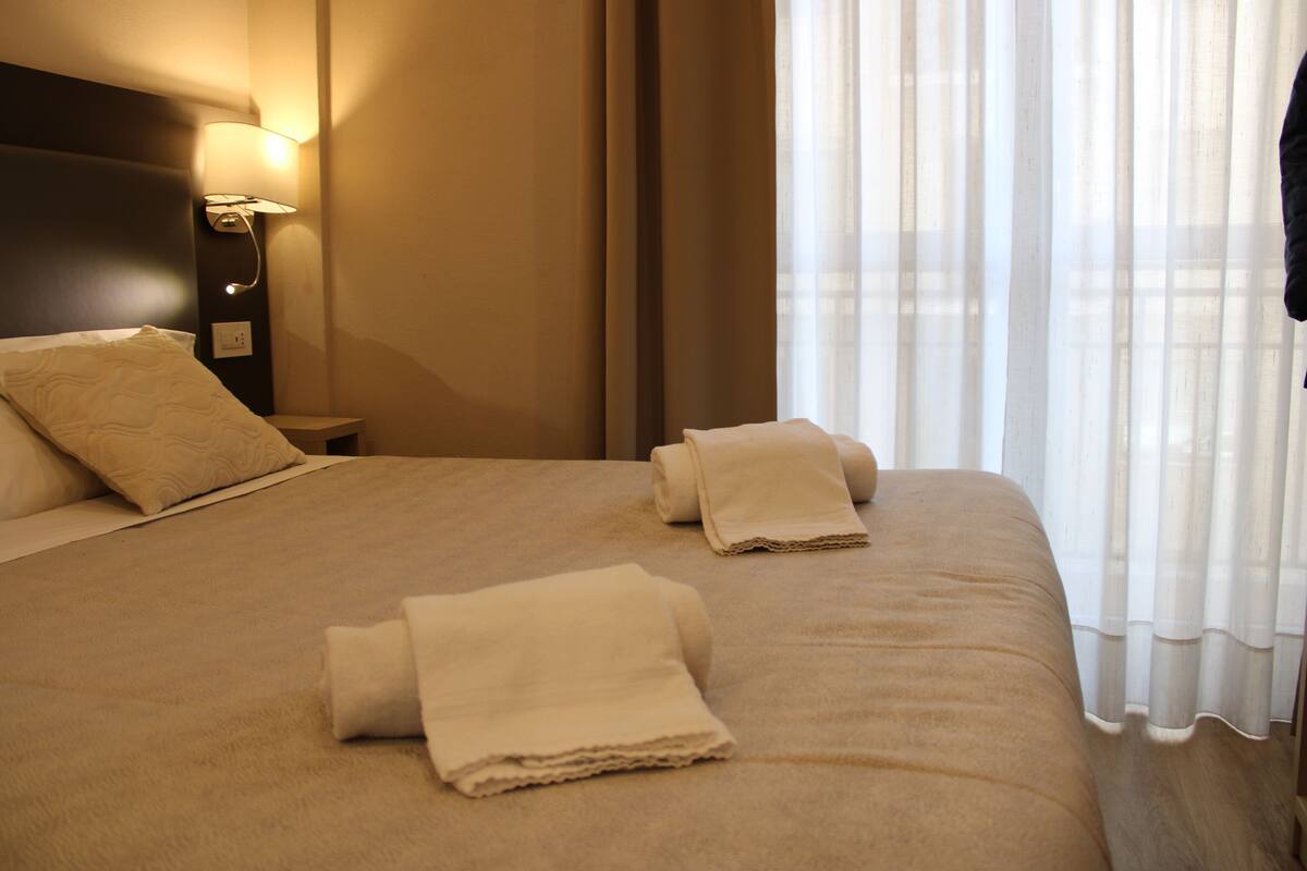 A neatly made bed with a headboard, a bedside lamp, and a window with sheer curtains.