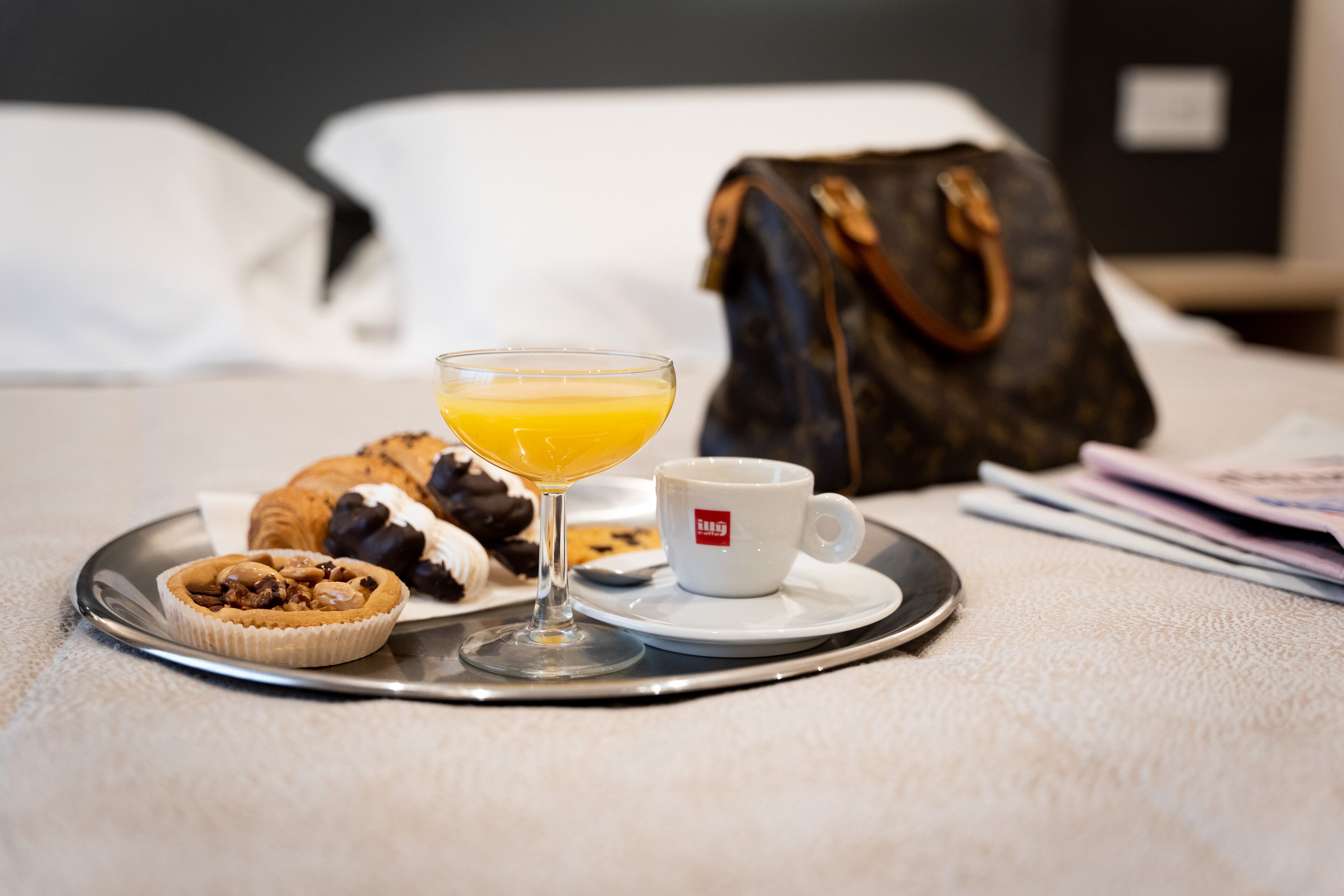 A breakfast tray with a croissant, a muffin, a glass of orange juice, and a cup of coffee on a hotel bed.