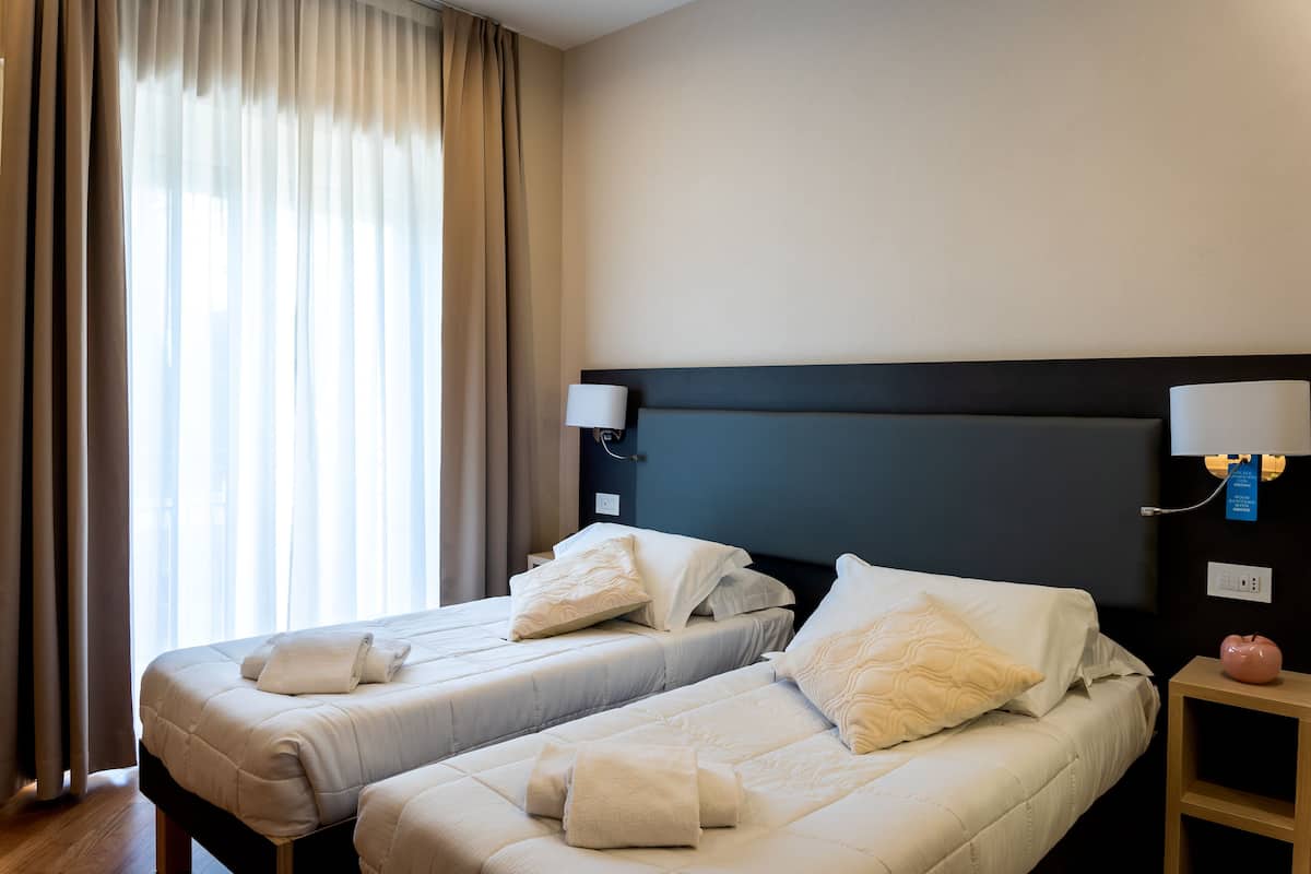 A hotel room with two beds, a nightstand with a lamp, and a view of the outdoors through sheer curtains.