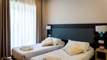 A hotel room with two beds, a nightstand with a lamp, and a view of the outdoors through sheer curtains.