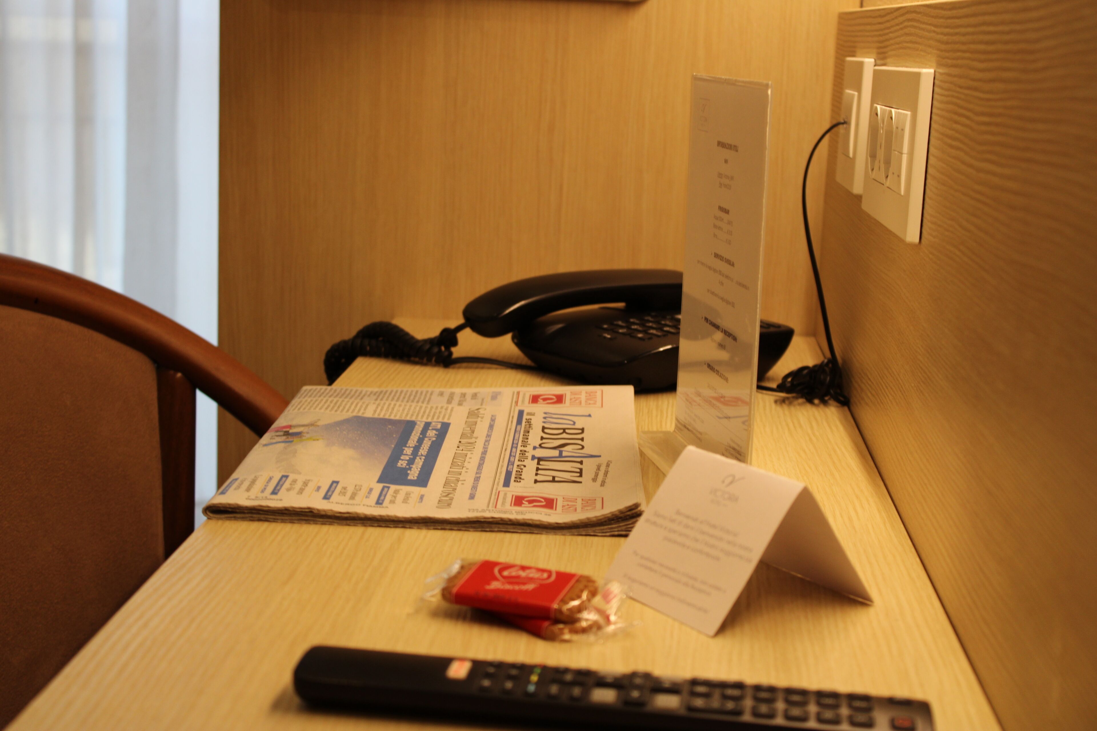 Escritorio de habitación de hotel con teléfono, control remoto, periódico y un pequeño paquete de snacks.