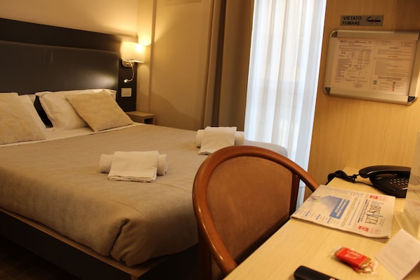 A hotel room with a bed, a bedside table with a lamp, a chair, a desk with a phone, and a bulletin board with information.