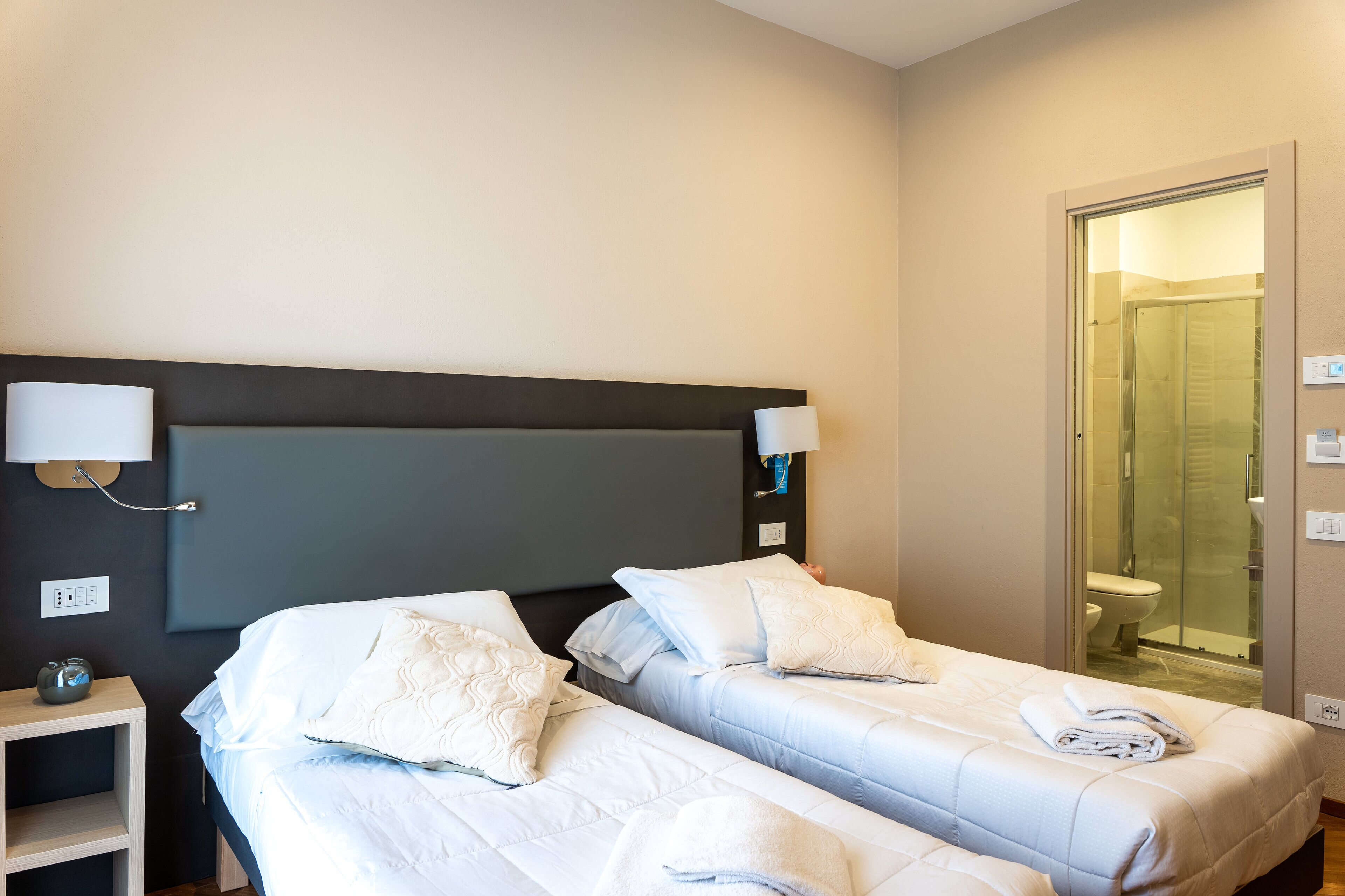Ein Hotelzimmer mit zwei Betten, einem Nachttisch, einer Wandleuchte und einer Duschkabine aus Glas.
