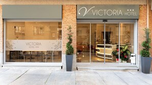 Exterior del Hotel Victoria, con amplios ventanales de vidrio, fachada de ladrillo y plantas en macetas.