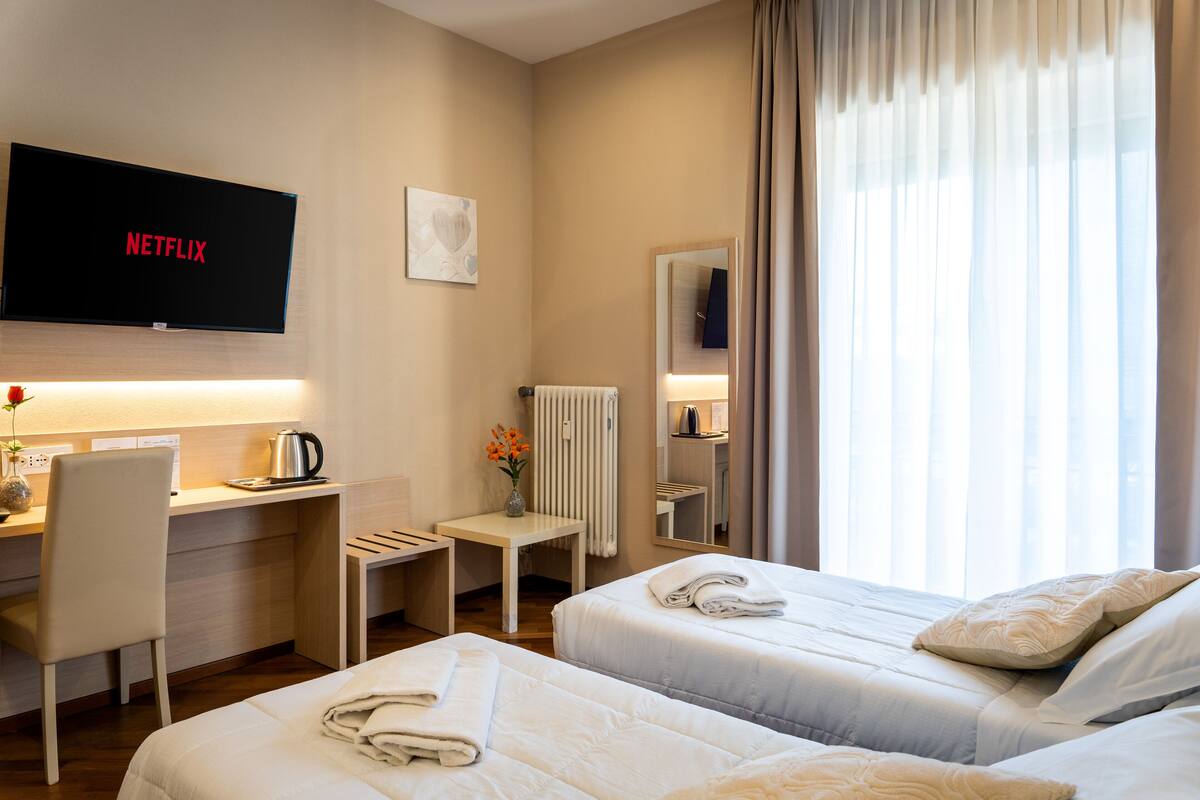 A hotel room with two beds, a desk, a TV displaying 'NETFLIX', a radiator, and a window with curtains.
