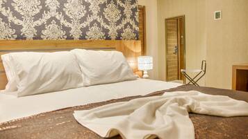 Quarto de hotel com cama, mesa de cabeceira com luminária, uma cadeira e uma porta.