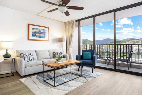 A modern living room with a sofa, coffee table, and a view of mountains through large windows. - Breezy condo w/ private lanai & beach access (Honolulu)