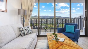 A modern living room with a sofa, a blue armchair, a wooden coffee table, and a view of mountains through large glass doors. - Breezy condo w/ private lanai & beach access (Honolulu)