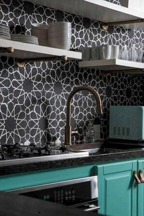 A kitchen with a geometric patterned backsplash, a stainless steel range hood, and teal cabinetry.