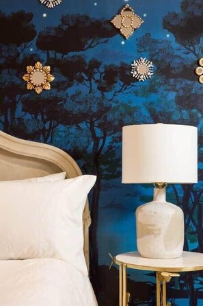 A bedroom with a bed, a white lamp on a small table, and a wall mural of trees and flowers.