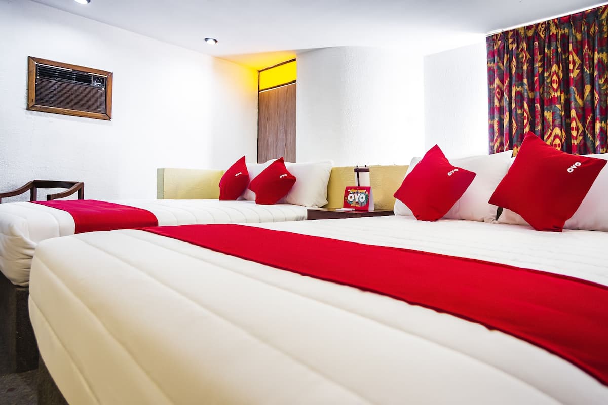 A hotel room with two beds, a bedside table with a red OYO branded item, a window with a wooden frame, and a colorful curtain.
