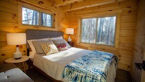 A wooden cabin bedroom with a bed, two bedside lamps, a window, and a view of trees. - Tanager cabin at Driftless Creek (Viroqua)