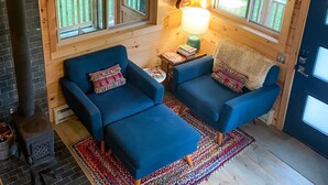 A cozy cabin interior with two blue armchairs, a wooden fireplace, and a large window overlooking a deck. - Tanager cabin at Driftless Creek (Viroqua)
