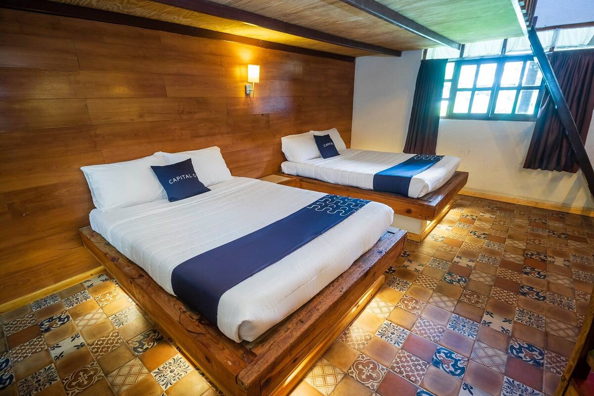 A hotel room with two beds, wooden headboards, a patterned floor, and a window with curtains.