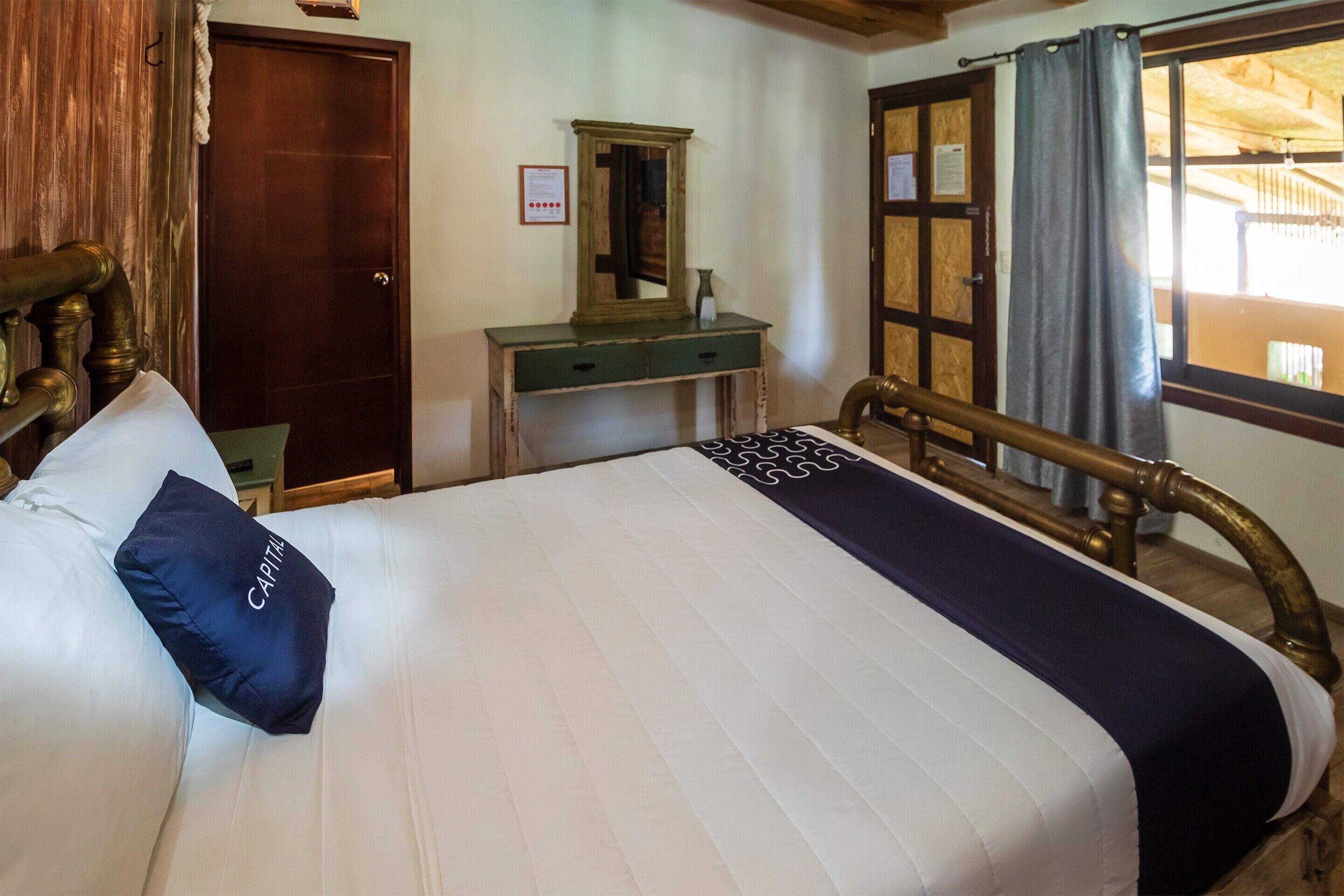 Una habitación de hotel con cama, cabecera de madera, escritorio con espejo y ventana con cortinas.