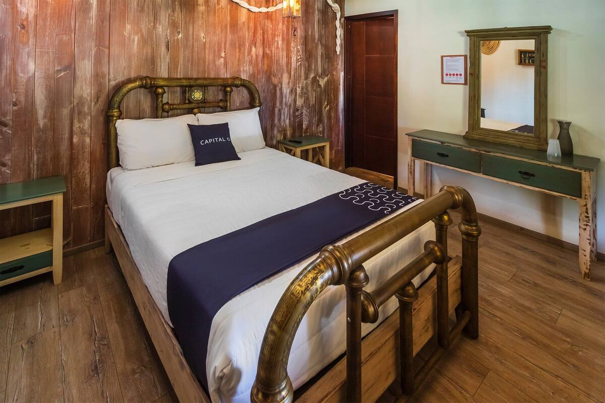 A wooden bed with a headboard, a bedside table, a dresser with a mirror, and a framed certificate on the wall.
