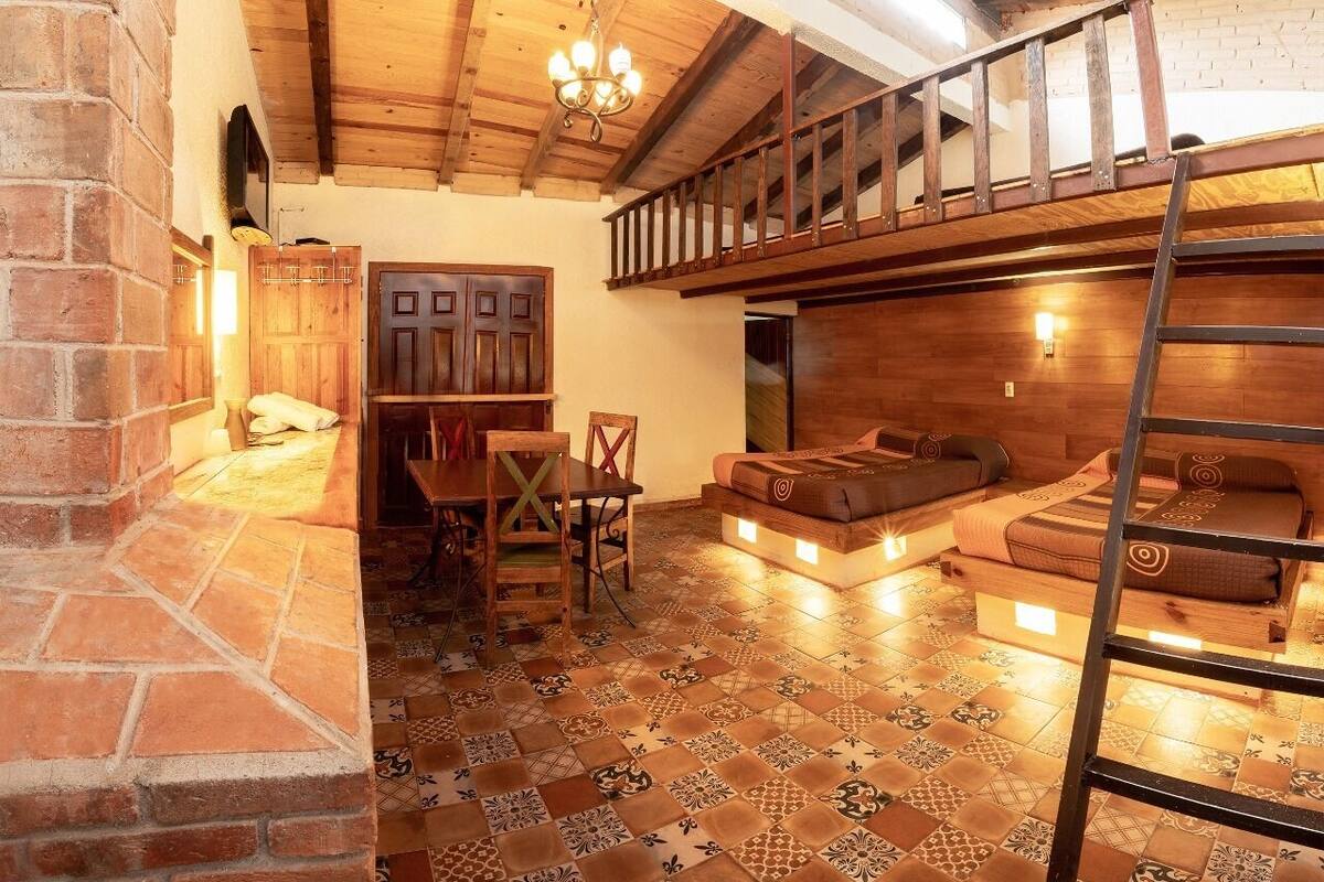 A cozy room with a wooden ceiling, a brick wall, a dining area with a table and chairs, a sofa, and a ladder.