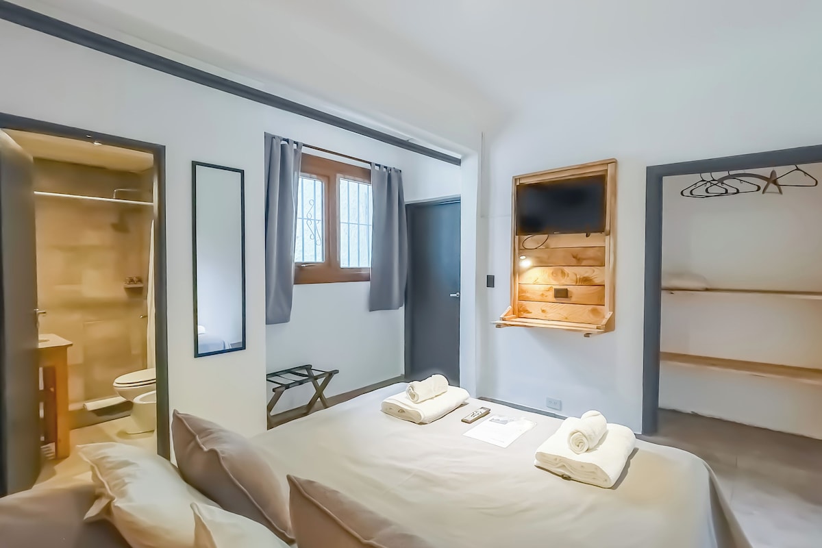 Une chambre d’hôtel comprenant un lit, une petite table, un miroir, une fenêtre avec des rideaux, une étagère en bois et une armoire.