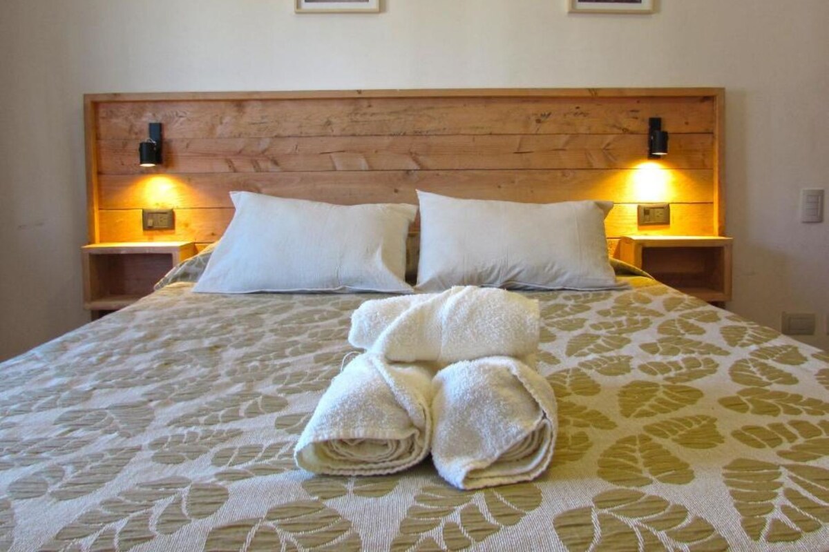 A neatly made bed with a patterned bedspread, two white pillows, and two rolled towels on the bed. There are two wall-mounted nightstands with lamps on either side of the bed.