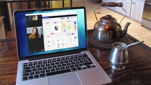 Ein Laptop auf einem Holztisch, zusammen mit einer Teekanne und einer Tasse, in einem Restaurant mit Holztischen und -stühlen, im Hintergrund eine Küchenzeile.
