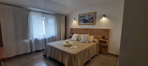 A hotel room with a wooden headboard, a bed with beige bedding, a bedside table, a nightstand, and a window with sheer curtains. - Huella Andina (San Carlos de Bariloche)