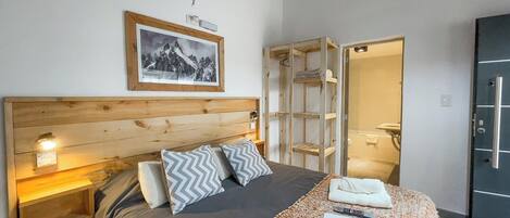 A bedroom with a wooden headboard, a bed with grey and white zigzag pillows, a framed picture on the wall, and a bathroom visible through an open door.