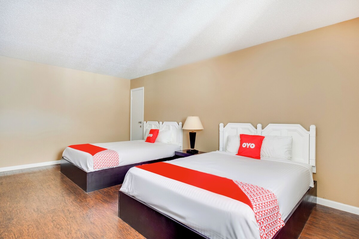 A hotel room with two beds, each with white bedding and red accents, a nightstand with a lamp, and a door with a red sign.