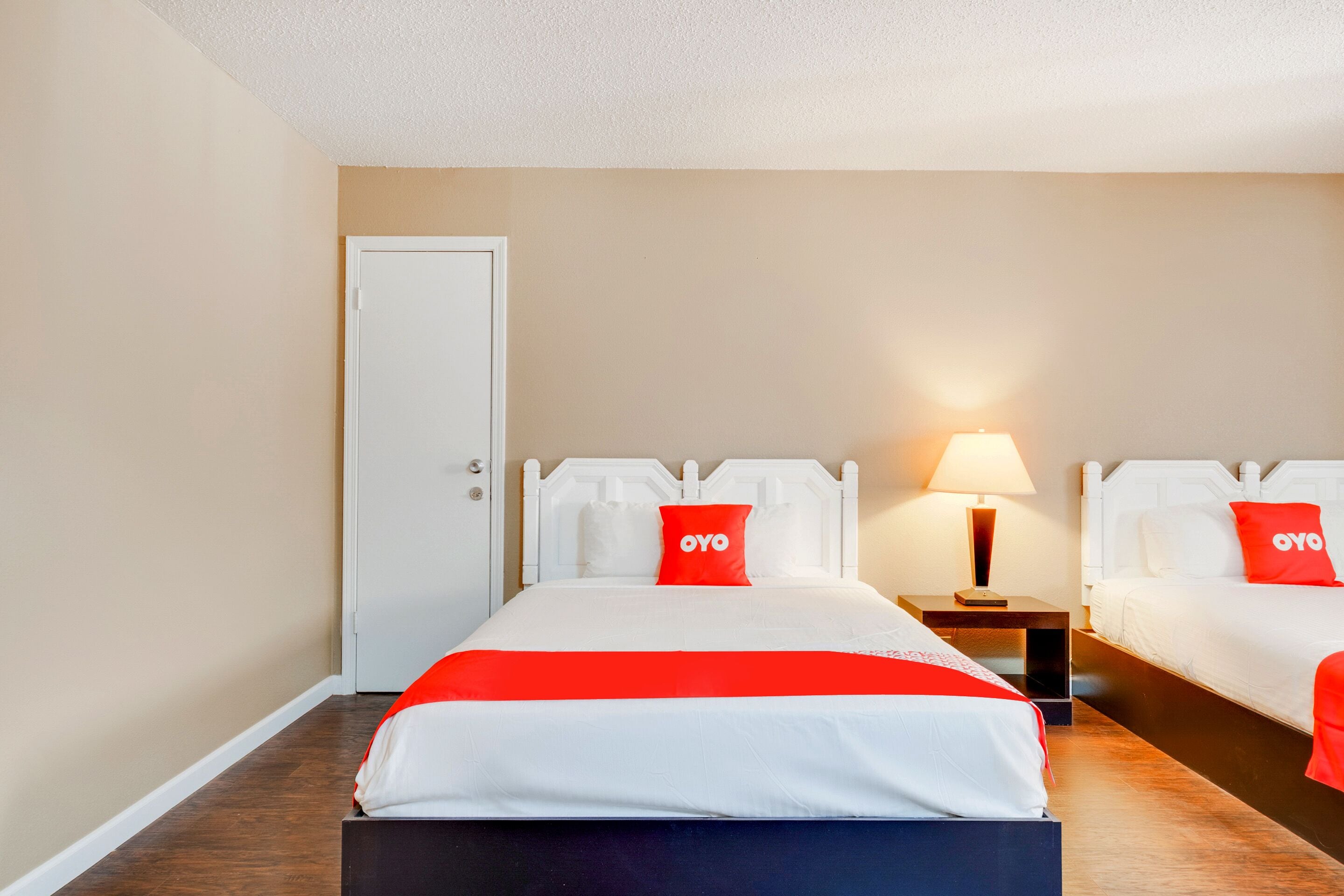 Een hotelkamer met twee bedden, een deur, een nachtkastje met een lamp en een rood-witte spreidek.