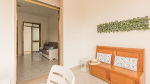 Een kamer met een houten bankje, een witte stoel en een kleine tafel met een kaars. Een deur leidt naar een andere kamer met een bank en een plant.