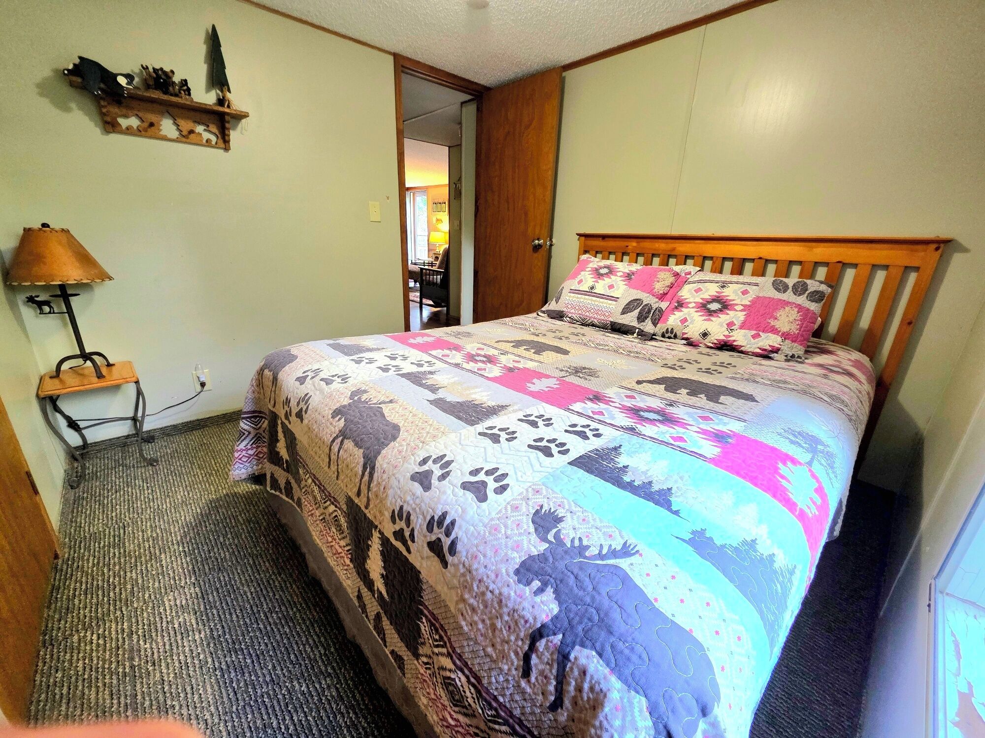 A bedroom with a bed, a nightstand, a lamp, and a wooden shelf with decorative items.
