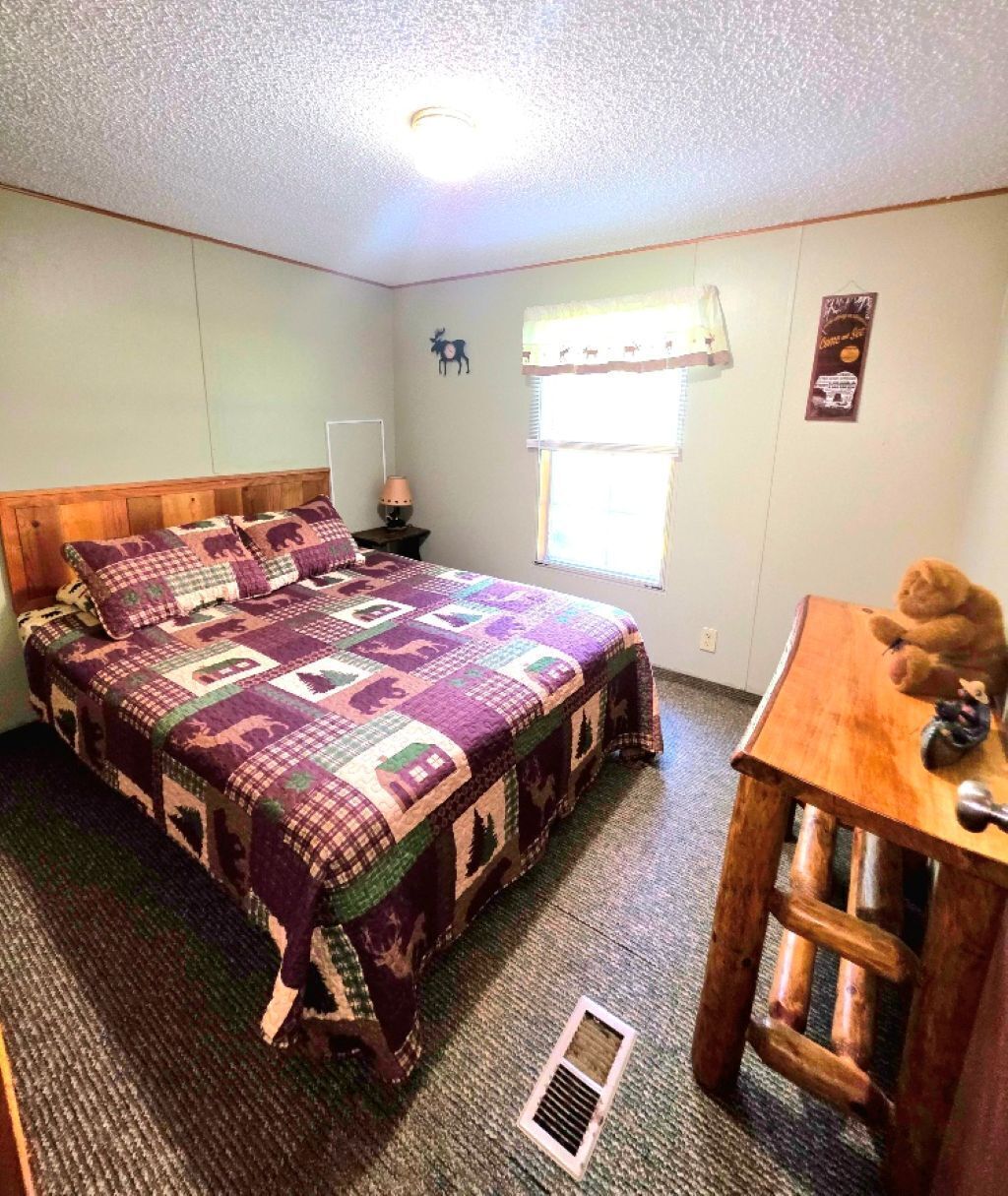 Quarto com cama de madeira, mesa de cabeceira, janela com cortina, um urso de pelúcia sobre uma mesa e um objeto decorativo na parede.