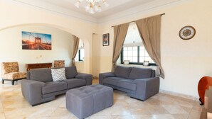 A spacious living room with two grey sofas, a matching ottoman, a wooden sideboard, and a large window with curtains. - Capital O Hotel San Jose, San Luis PotosÃ­ (San Luis Potosi)