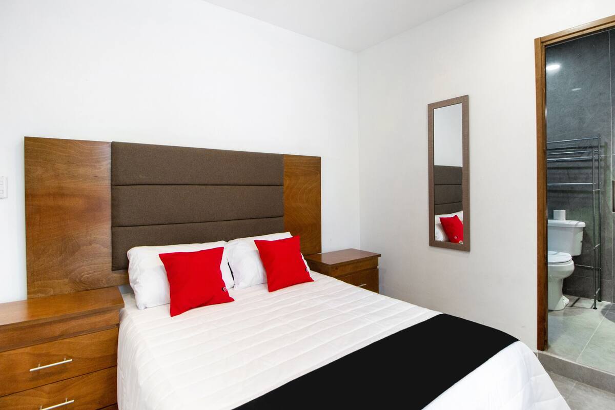 Una cama bien hecha con almohadas blancas y rojas, un cabecero de madera y una mesita de noche de madera con cajones.
